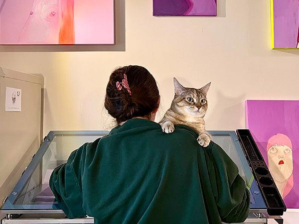 Ambivalently Yours draws at her desk with her cat on her shoulder